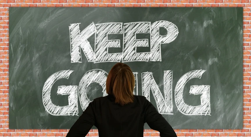 a woman standing in front of a chalkboard that says keep going, by Jeka Kemp, trending on pixabay, happening, -step 50, resignation, painted on a brick wall, interrupting the big game