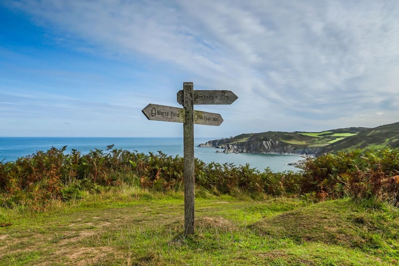 a wooden sign sitting on top of a lush green hillside, by Edward Corbett, shutterstock, looking out over the sea, devon cady-lee, round about to start, leading to a beautiful