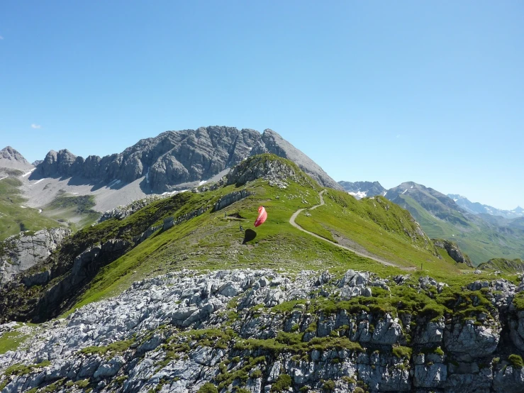 a man flying a kite on top of a lush green hillside, a picture, by Werner Andermatt, flickr, dolomites in background, balloon, walking towards the camera, tent