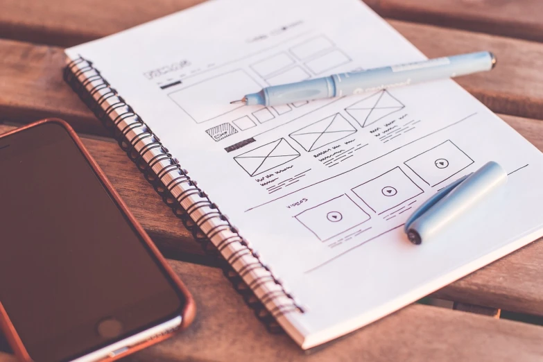 a notebook sitting on top of a wooden table next to a cell phone, a wireframe diagram, by Matt Cavotta, pexels, designer pencil sketch, slick elegant design, ad image, thumbnail
