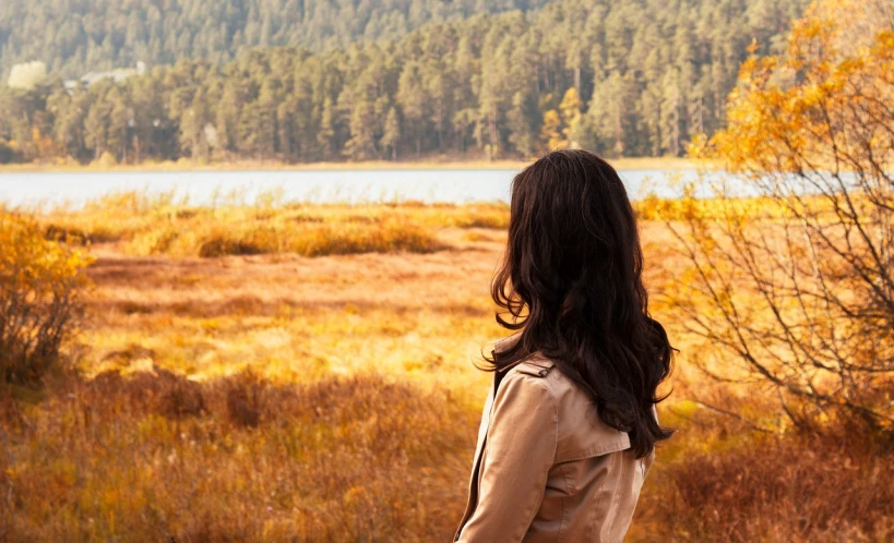 a woman standing in a field with trees in the background, inspired by Andrew Wyeth, pexels, near lake baikal, autum, relaxed. gold background, view from back