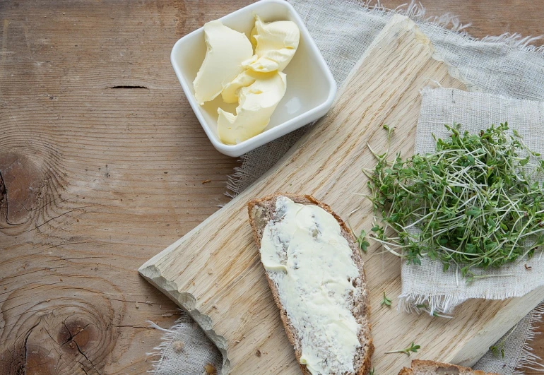 a piece of bread sitting on top of a wooden cutting board, a picture, by Dietmar Damerau, shutterstock, mayonnaise, cream and white color scheme, clover, butter