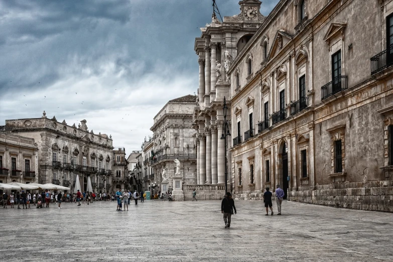 a group of people walking down a street next to tall buildings, by Mirabello Cavalori, shutterstock, neoclassicism, conversano, a wide open courtyard in an epic, on a cloudy day, stock photo