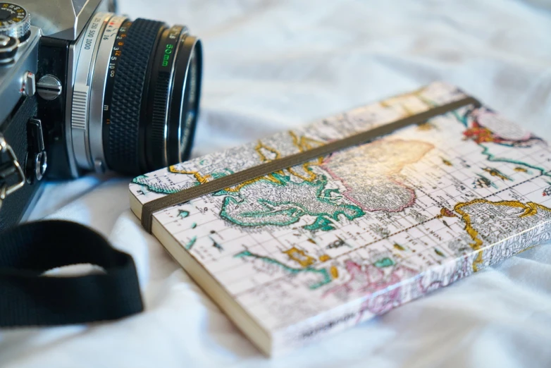 a camera and a notebook on a bed, a picture, by Caro Niederer, pexels, visual art, cartography map art, marble!! (eos 5ds r, 🚿🗝📝, beauty shot