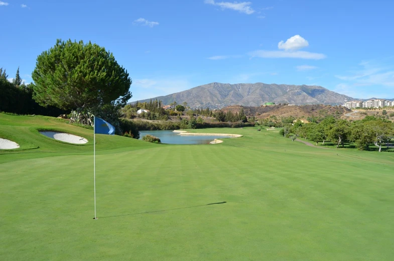 a golf course with a lake and mountains in the background, a picture, by Robbie Trevino, marbella landscape, green flag, back facing, with slight stubble