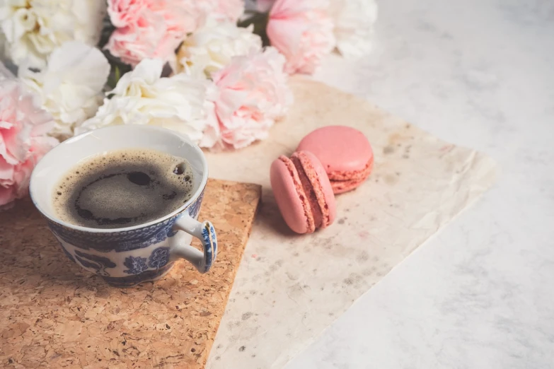 a cup of coffee and a macaron on a table, a picture, by Emma Andijewska, shutterstock contest winner, romanticism, pink flowers, food styling, pink and blue colour, on old parchment paper