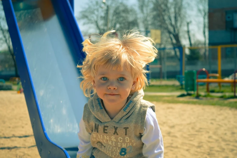 a little girl that is standing in front of a slide, a portrait, by Jan Rustem, pexels, blonde shaggy hair, boy with neutral face, at the park on a beautiful day, disheveled