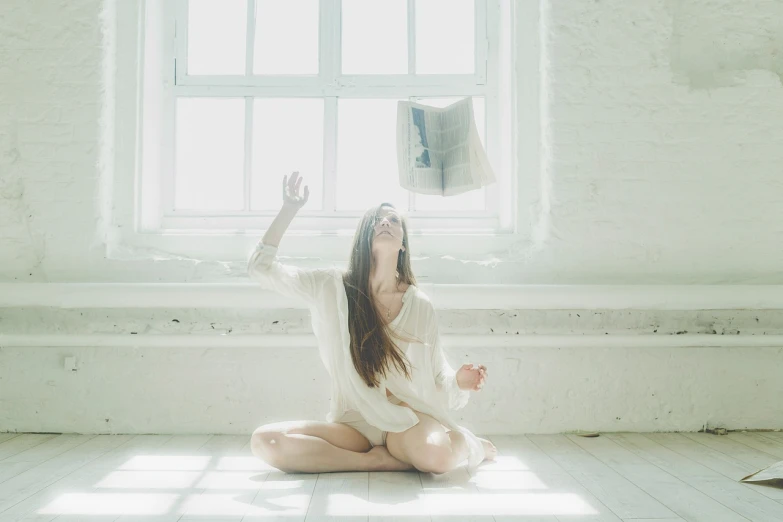a woman sitting on the floor in front of a window, a picture, inspired by Anka Zhuravleva, tumblr, light and space, with arms up, holding a tattered magical book, white studio, the girl and the sun