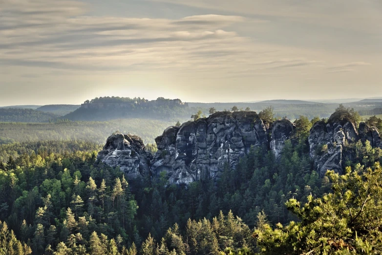 a group of rocks sitting on top of a lush green forest, a picture, by Karl Hagedorn, morning sunrise, high cliff, lower saxony, distant photo