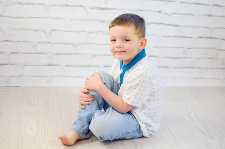 a young boy sitting on the floor in front of a brick wall, a picture, figuration libre, white shirt and blue jeans, wearing collar on neck, 8 k photo