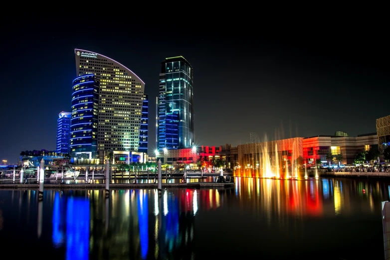 a city at night with a fountain in the foreground, shutterstock, hurufiyya, layers of colorful reflections, dubai, city docks, glass buildings