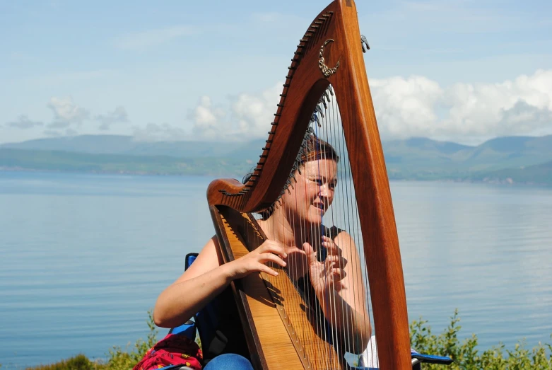 a woman playing a harp by the water, a portrait, by Alison Watt, flickr, cloudless-crear-sky, with mountains in background, very very happy, festival