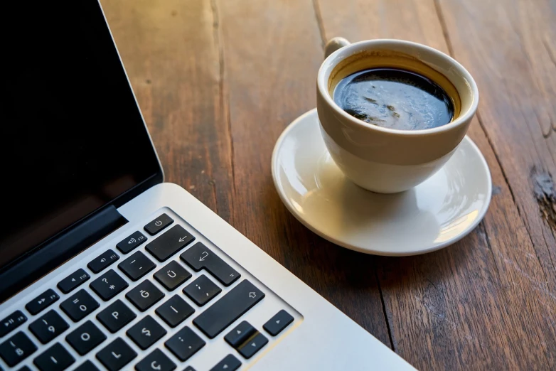 a cup of coffee on a saucer next to a laptop, shutterstock, stock photo, closeup portrait shot, on wooden table, high res photo