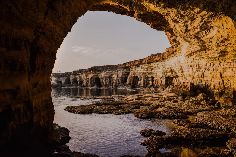 a cave filled with lots of rocks next to a body of water, a picture, by Simon Marmion, pexels contest winner, les nabis, cyprus, archway, morning glow, layers of strata