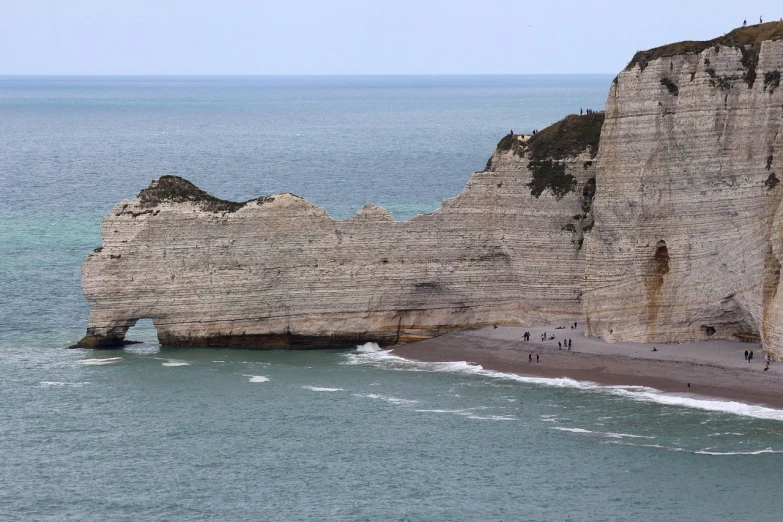 a group of people standing on top of a beach next to the ocean, a picture, by Raphaël Collin, chalk cliffs above, massive arch, wikimedia commons, geological strata