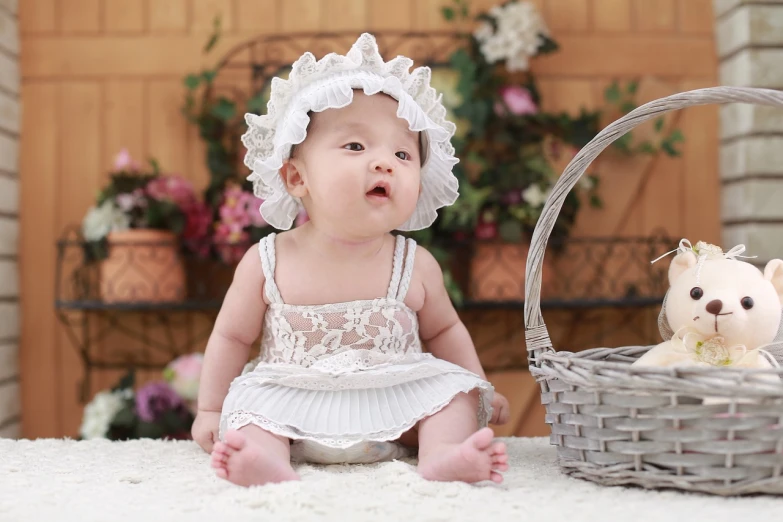 a baby sitting next to a basket with a teddy bear, a picture, pexels, happening, dressed in a frilly ((lace)), traditional chinese, wearing a white bathing cap, beautiful girlfriend