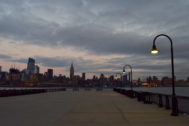 a bench sitting on top of a sidewalk next to a body of water, a picture, by Joseph Pisani, unsplash, on liberty island, overcast dusk, dusk on a city street, video still