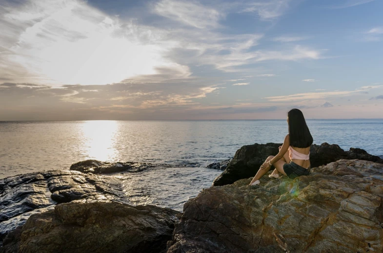 a woman sitting on top of a rock next to the ocean, a picture, girl watching sunset, wide shot photo, stoic and calm, full res