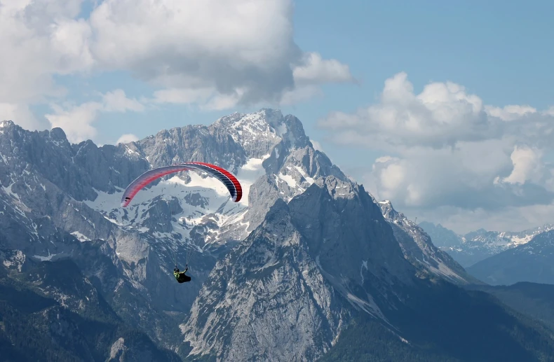 a person paragliding in the mountains on a sunny day, a picture, figuration libre, rocky mountains in background, sturm und drang, flying vehicles, viewed from a distance