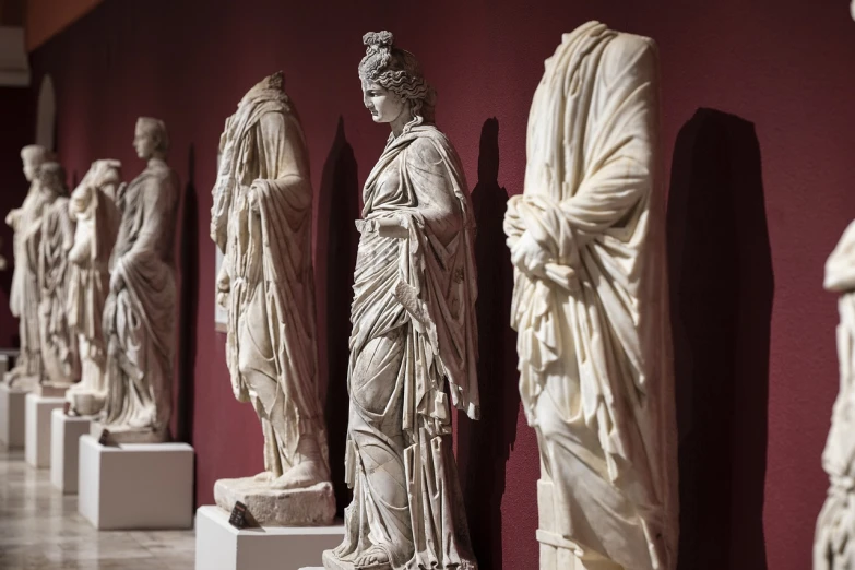 a row of statues in front of a red wall, a marble sculpture, inspired by Pietro Testa, shutterstock, metropolitan museum collection, lady of elche, female, roman emperor