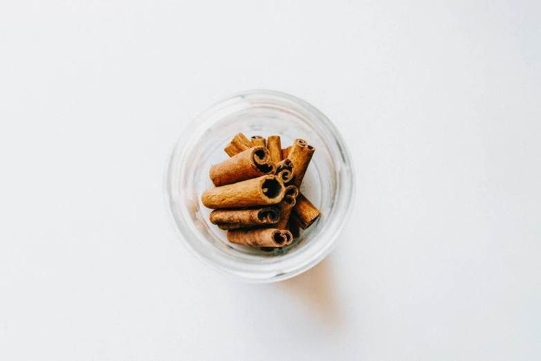 a glass filled with cinnamon sticks on top of a white table, by Emma Andijewska, view from above, glass jar, pepper, adorable