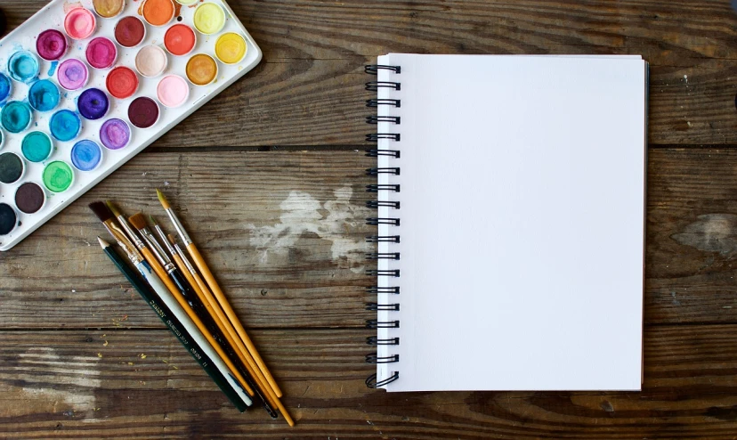 an open notebook sitting on top of a wooden table, a drawing, inspired by artist, pexels, process art, white paint, paintbrush and palettes, background image, no text!
