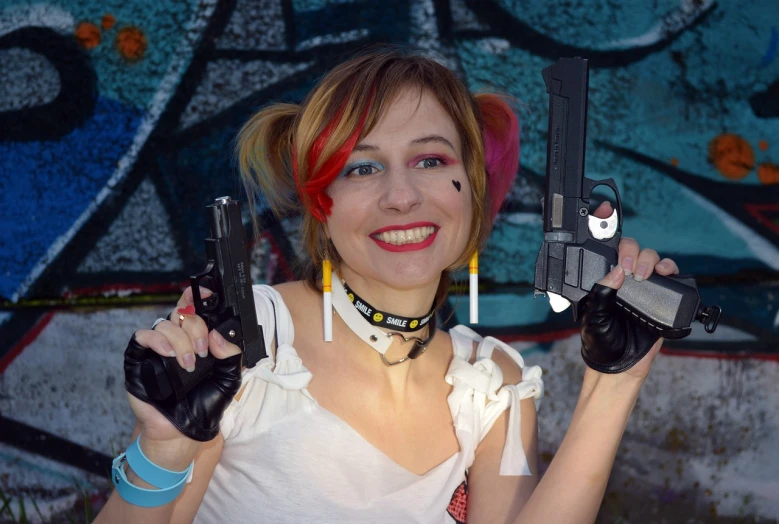 a woman holding two guns in front of a graffiti wall, a photo, inspired by Earle Bergey, deviantart, portrait of harley quinn, 1 / 4 headshot, rena nounen style 3/4, she is happy