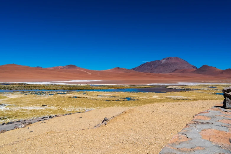 a man standing on top of a sandy beach next to a body of water, a photo, by Niklaus Manuel, shutterstock, color field, andes, near a small lake, sparse frozen landscape, extreme panoramic