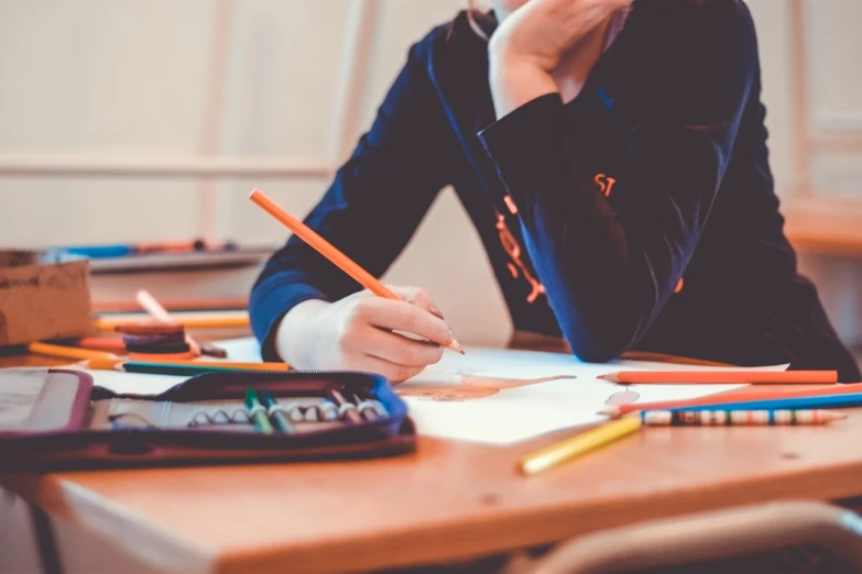 a woman sitting at a desk writing on a piece of paper, a child's drawing, by Joseph-Marie Vien, pexels, school uniform, orange and blue colors, holding pencil, 4 0 9 6