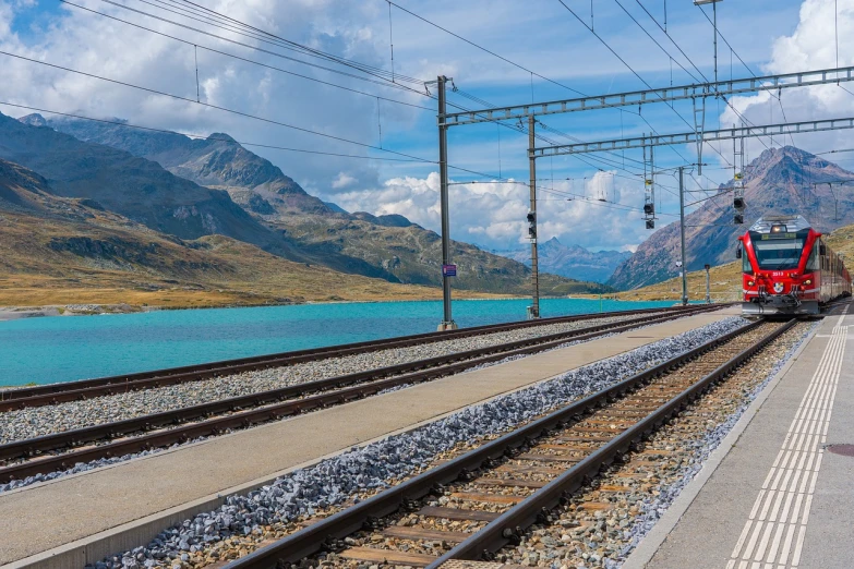 a red train traveling down train tracks next to a body of water, a stock photo, by Cedric Peyravernay, shutterstock, brown and cyan blue color scheme, swiss, electric cables, train station in summer