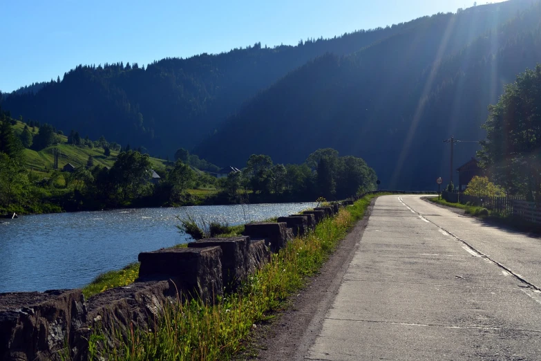 a man riding a bike down a road next to a river, a picture, by Karl Pümpin, flickr, les nabis, black forest, sunny morning, road between hills, near a jetty