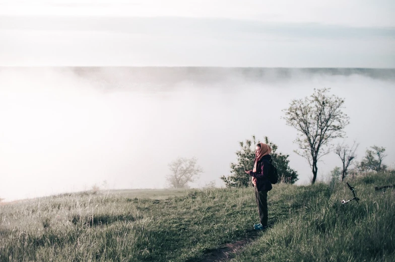 a man standing on top of a grass covered hillside, a picture, by Matthias Weischer, pexels, romanticism, fog on the water, girl looks at the space, ukraine. photography, low quality footage