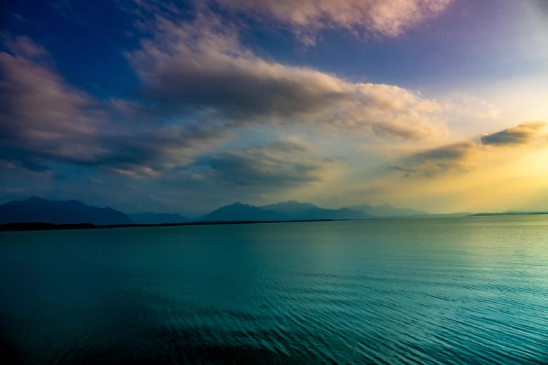 a large body of water under a cloudy sky, by Michalis Oikonomou, calm vivid colors, distant mountains lights photo, very beautiful photo, green and blue colors