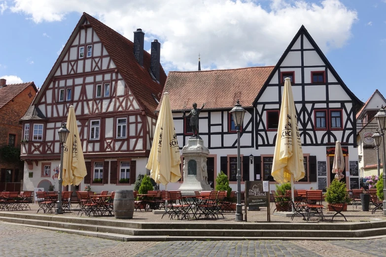 a group of tables and umbrellas in front of a building, a portrait, by Sigmund Freudenberger, pexels, timbered house with bricks, monument, in dunwall, white buildings with red roofs