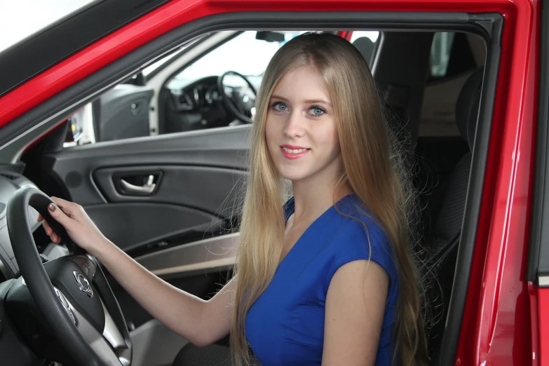 a woman sitting in the driver's seat of a red car, long blonde hair and blue eyes, nadezhda tikhomirova, new vehicle, detailed information