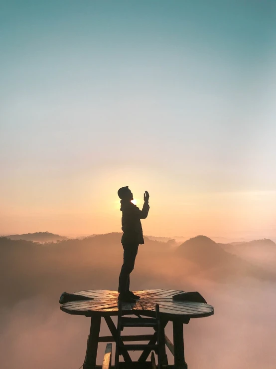 a man standing on top of a table on top of a mountain, a picture, pexels, romanticism, indistinct man with his hand up, phone wallpaper, glowing halo above his head, isolated