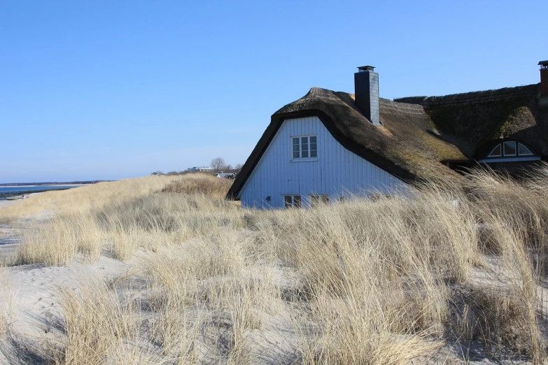 a white house sitting on top of a sandy beach, by Christen Købke, pixabay, dry grass, simple gable roofs, location of a dark old house, alternate angle