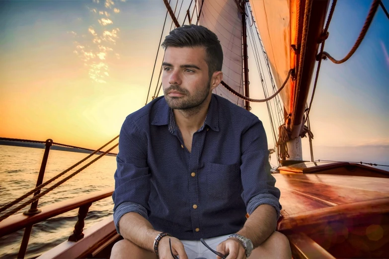 a man in a blue shirt sitting on a boat, a portrait, by Juan O'Gorman, pixabay contest winner, hurufiyya, zachary quinto, 26 year old man on a sailboat, sunset in the background, professional profile picture