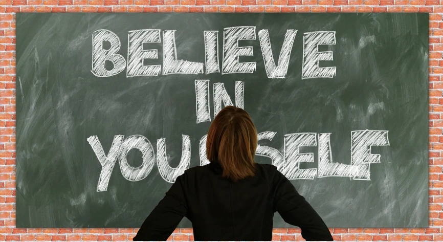 a woman standing in front of a blackboard with believe in yourself written on it, a picture, inspired by John Maxwell, pixabay, happening, barbara kruger, standing in front of a mirror, looking upwards, person in foreground
