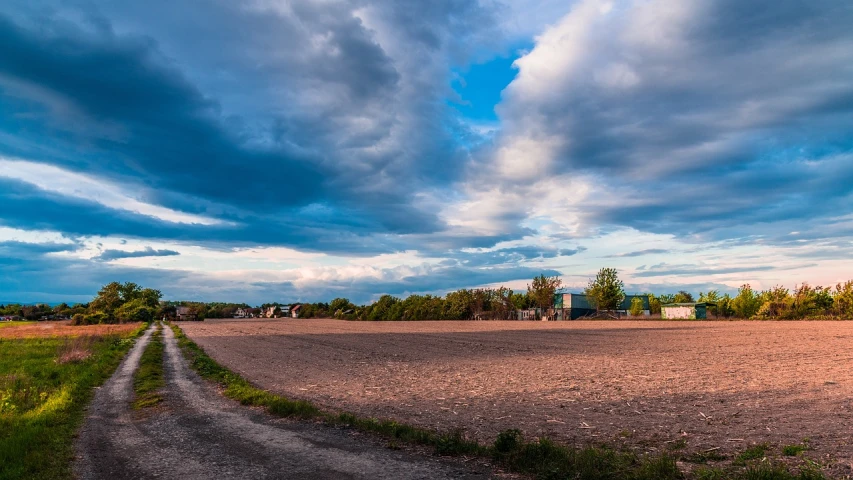 a dirt road next to a field under a cloudy sky, a portrait, by Karl Hagedorn, flickr, sunset panorama, small town surrounding, 1/1250s at f/2.8, nice spring afternoon lighting