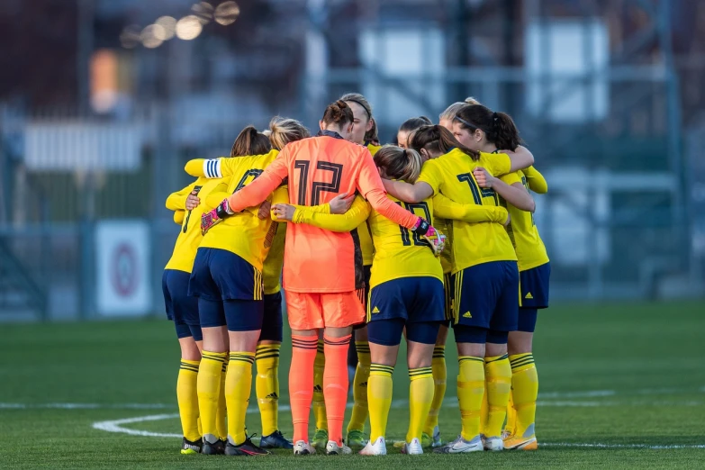 a group of women standing on top of a soccer field, a picture, inspired by Christen Dalsgaard, shutterstock, with yellow cloths, scotland, indoor shot, hugging