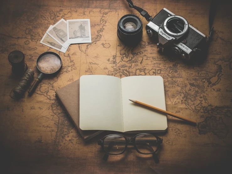 an open book sitting on top of a table next to a camera, a picture, map cartography, reading glasses, brown, sketching