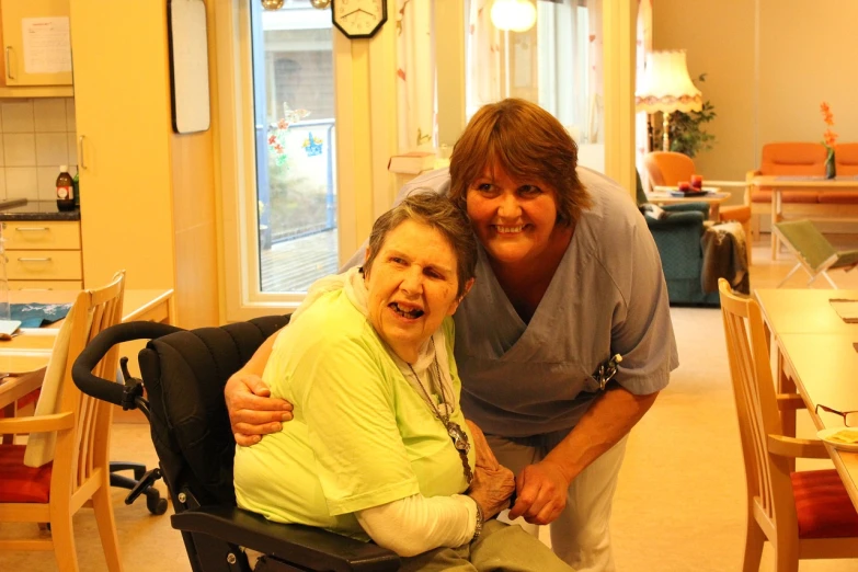 a couple of women standing next to each other in a room, a photo, by Pat Adams, cg society contest winner, sitting in a wheelchair, happy!!!, hug, bella