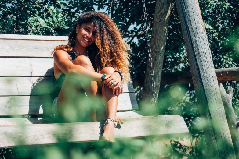 a woman sitting on top of a wooden bench, a portrait, pexels, happening, super cute funky black girl, green and brown tones, summer feeling, with long curly