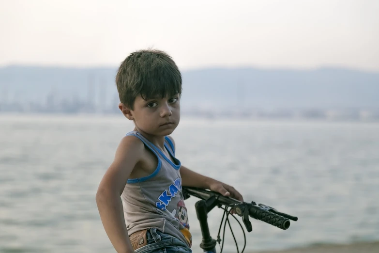 a little boy that is standing with a bike, by Ibrahim Kodra, at the waterside, an indifferent face, hayao miyazak, closeup portrait shot