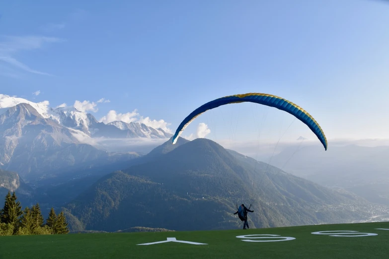 a man standing on top of a lush green field, a picture, flickr, parachutes, chamonix, early in the morning, gliding