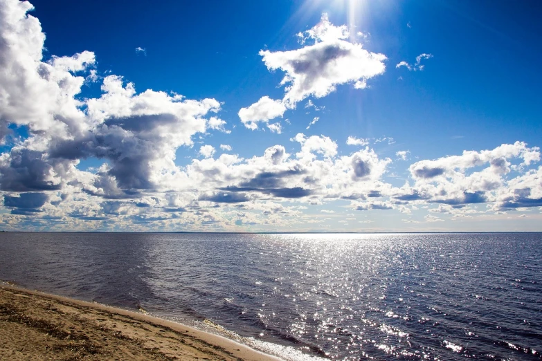 a large body of water next to a sandy beach, by Maksimilijan Vanka, pexels, among heavenly sunlit clouds, siberia!!, sunbeams blue sky, shiny silver