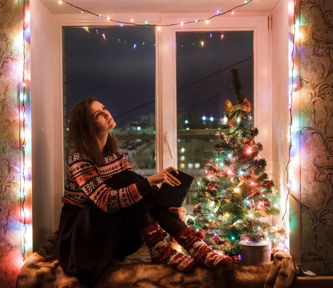 a woman sitting on a window sill in front of a christmas tree, a portrait, shutterstock, christmas lights, alexey gurylev, full length shot, wearing an oversized sweater