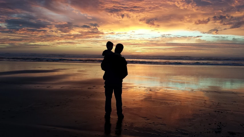 a couple of people standing on top of a sandy beach, a picture, by Matt Stewart, pixabay, father with child, lit in a dawn light, over his shoulder, son