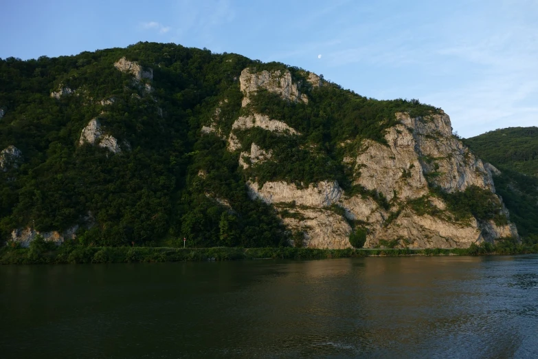 a large body of water with a mountain in the background, a picture, flickr, danube school, cliff side at dusk, with lots of vegetation, limestone, evening sun